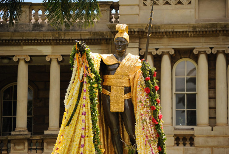 Stately bronze statue of King Kamehameha I, situated in the grounds of the Iolani Palace in Honolulu, Oahu, Hawaii. The impressive statue portrays King Kamehameha I standing tall and resolute, adorned in a magnificent cape woven with intricate gold designs. His left hand rests on his hip while his right arm extends outward, symbolizing his wise and visionary leadership. The statue is surrounded by lush greenery and set against the backdrop of the historic Iolani Palace, representing the enduring legacy of King Kamehameha I as the founding monarch of the Kingdom of Hawaii.