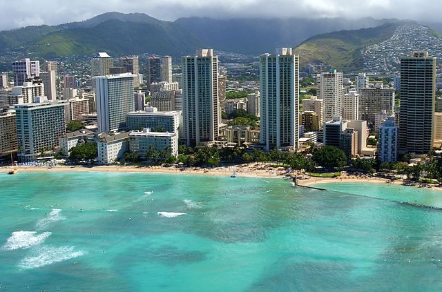 An aerial view of Waikiki Beach in Honolulu, Hawaii. The golden sandy beach stretches along the sparkling blue waters of the Pacific Ocean. Sunbathers relax under colorful umbrellas while surfers catch waves in the distance. Tall buildings line the shore, creating a vibrant cityscape backdrop. The palm tree-dotted shoreline and clear skies complete the picture-perfect scene of this iconic Hawaiian destination.